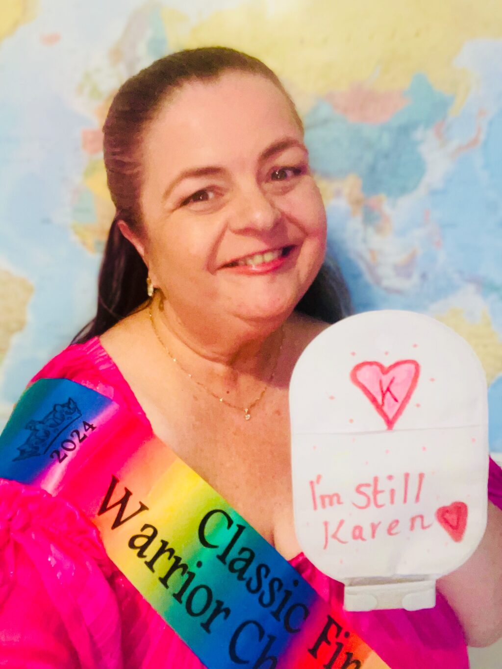 Photograph of a woman smiling wearing a pink shirt and rainbow sash with the words Classic and Warrior visible, she holds up a stoma bag with a red heart on it and the words “I’m still Karen”.